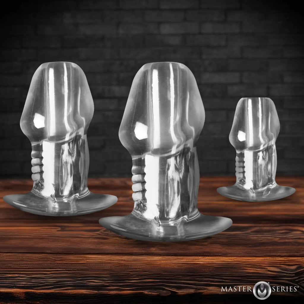 Master Series 3pc clear hollow anal plug set featuring phallic-shaped designs on a wooden surface against a brick wall.