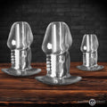 Master Series 3pc clear hollow anal plug set featuring phallic-shaped designs on a wooden surface against a brick wall.