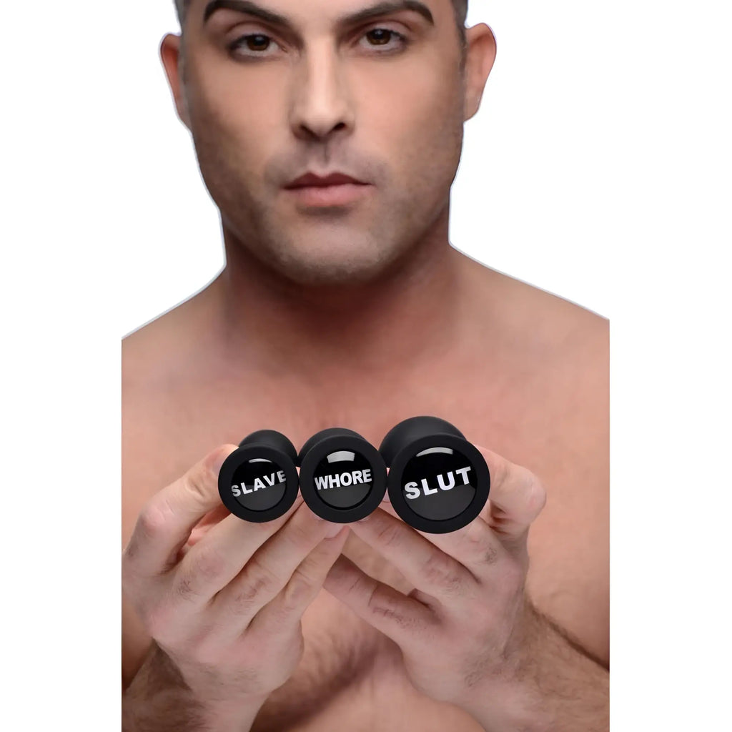 A man holding three black Master Series anal plugs of increasing sizes, each labeled with white text: SLAVE, WHORE, SLUT.