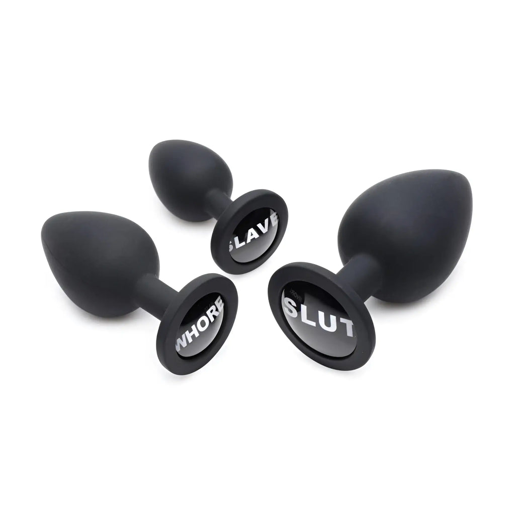 Three black Master Series Dirty Words anal plugs in varying sizes with SLAVE, WHORE, and SLUT printed on their bases.
