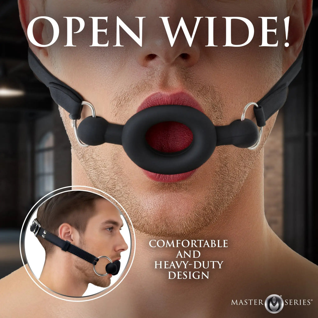 Master Series Forced Silicone Feeding Gag: Black ring-style mouth gag with adjustable straps worn by a male model.
