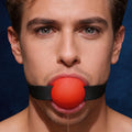 A man wearing a Master Series Gag Order Extreme ball gag, featuring a bright red silicone ball and black strap.
