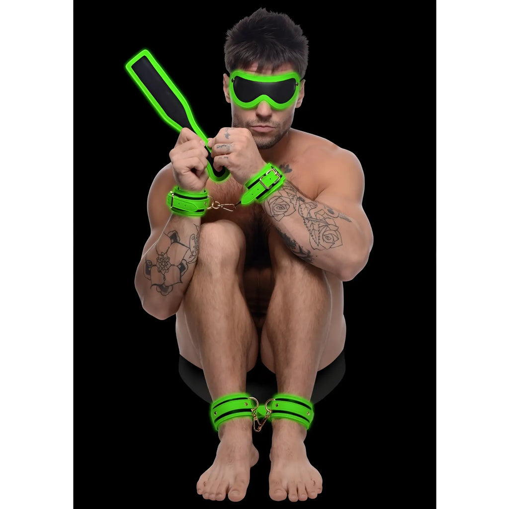 Master Series Kink in the Dark set: neon green cuffs, blindfold, and paddle worn by a tattooed man on black background.