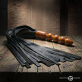 Master Series Lasher leather flogger with black multi-strand tails and a contoured wooden handle on a wooden table.