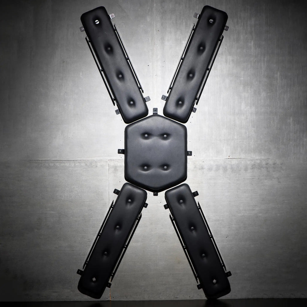 Master Series Masters St. Andrew's Cross: A wall-mounted black tufted leather X-shaped restraint against a gray wall.