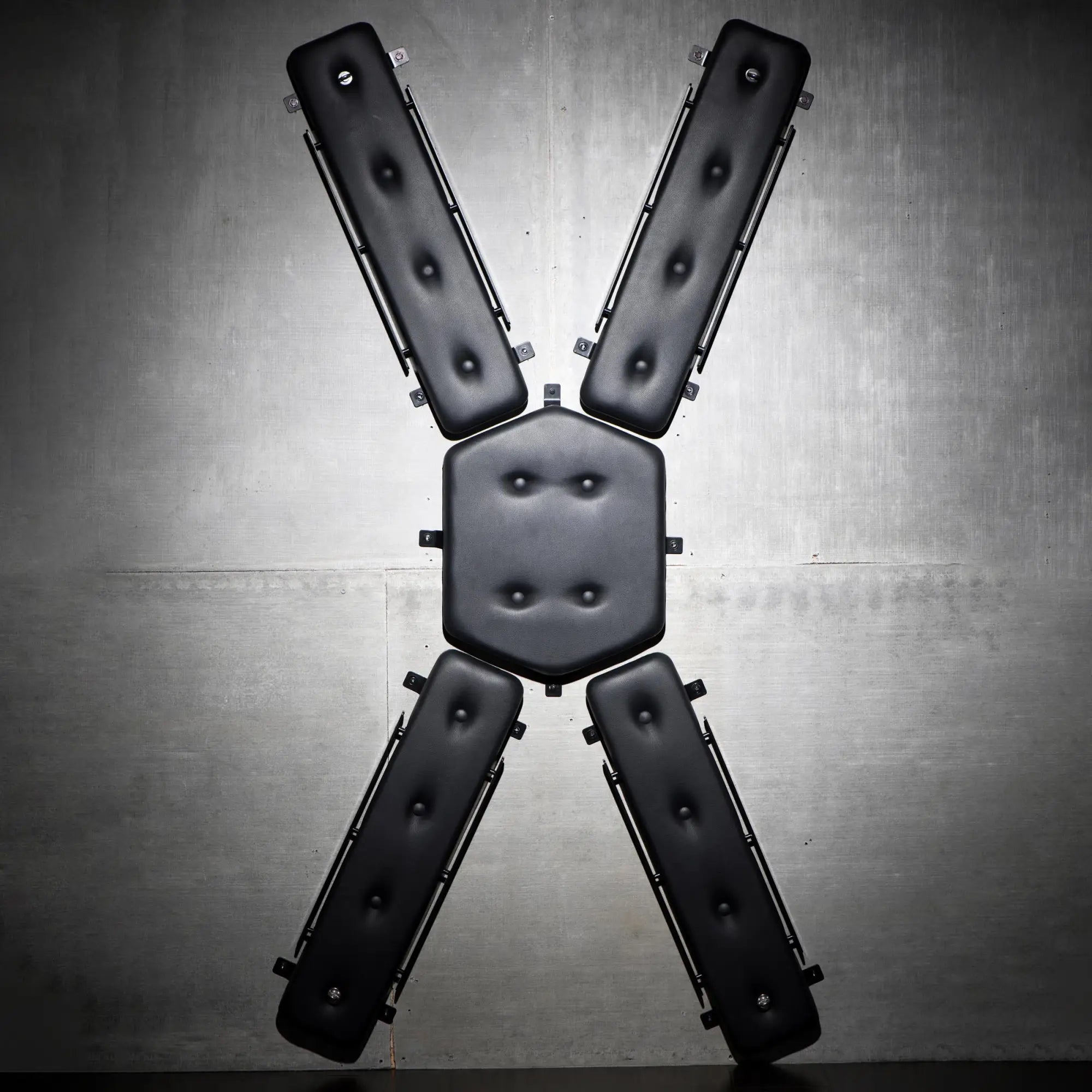 Master Series Masters St. Andrew's Cross: A wall-mounted black tufted leather X-shaped restraint against a gray wall.