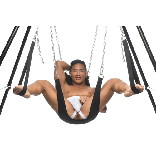 A smiling man sits in a black Master Series Nylon Sex Sling suspended by metal chains on a black metal frame.