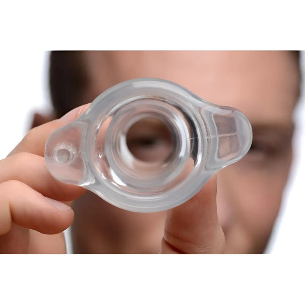 Master Series PeepHole clear, hollow silicone anal plug held up to the camera, looking through its center opening.