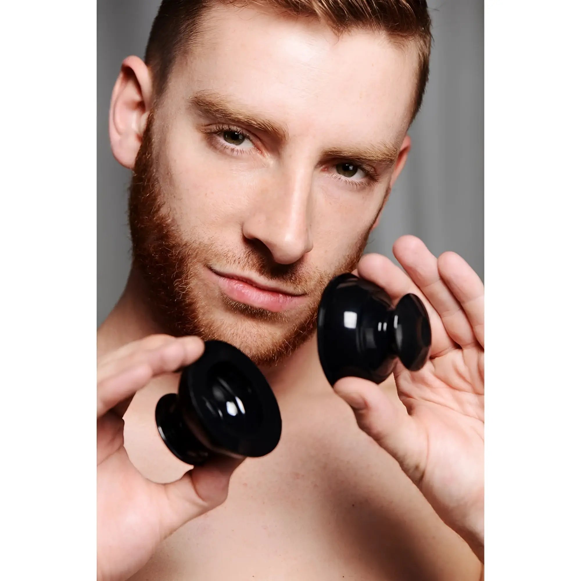A man holding two black, plunger-style Master Series silicone nipple suckers in front of his chest.