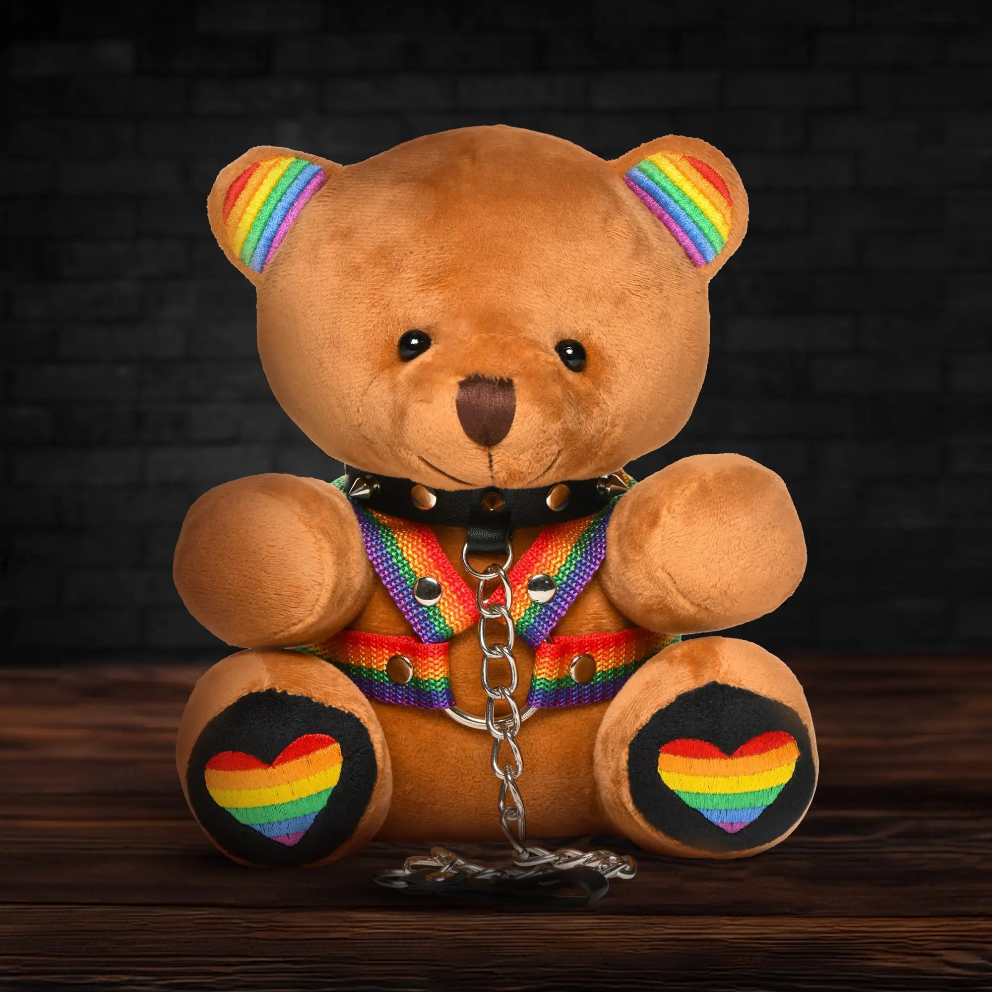 Master Series Pride Bear: a brown teddy bear wearing a spiked collar, rainbow harness, and chain on a wooden surface.