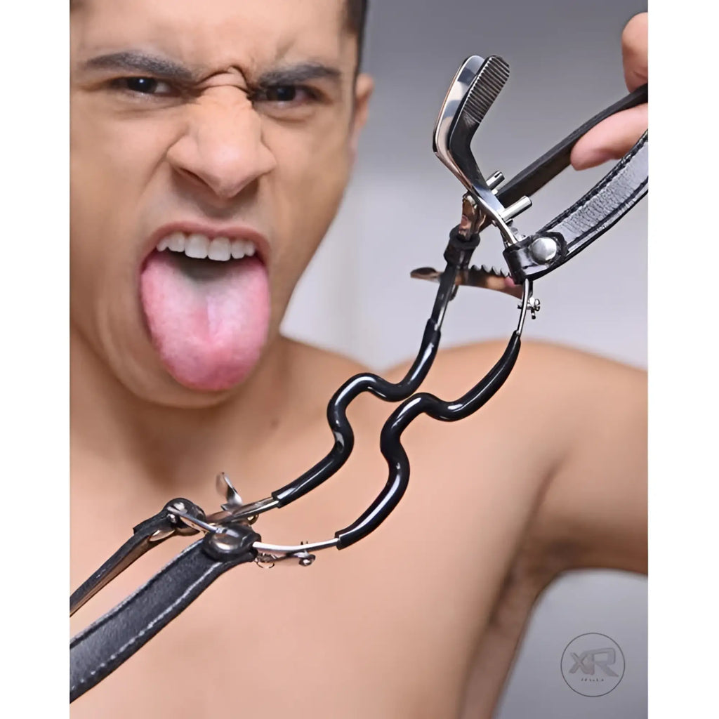 Master Series ratchet-style Jennings mouth gag with black straps held near the face of a person with tongue out.