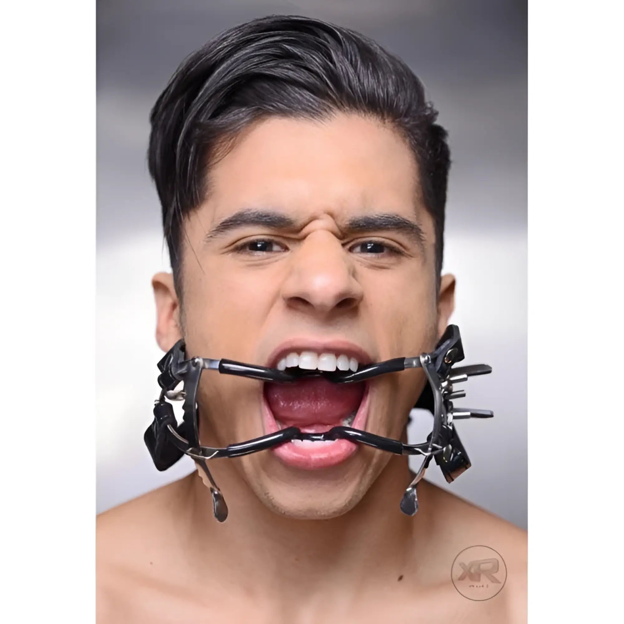 A person wearing a black Master Series ratchet style Jennings mouth gag with a head strap, holding their mouth open.