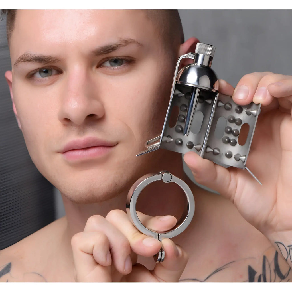 A man holds a silver Master Series Spiked Chamber Chastity Cage with an open, spiked interior and a separate base ring.