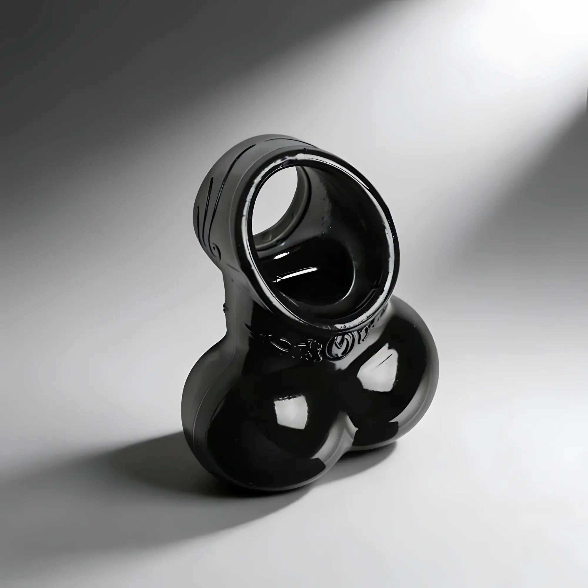 Master Series Squeeze My Sack: A shiny black, ergonomic silicone erection enhancer and scrotum pouch on a grey background.