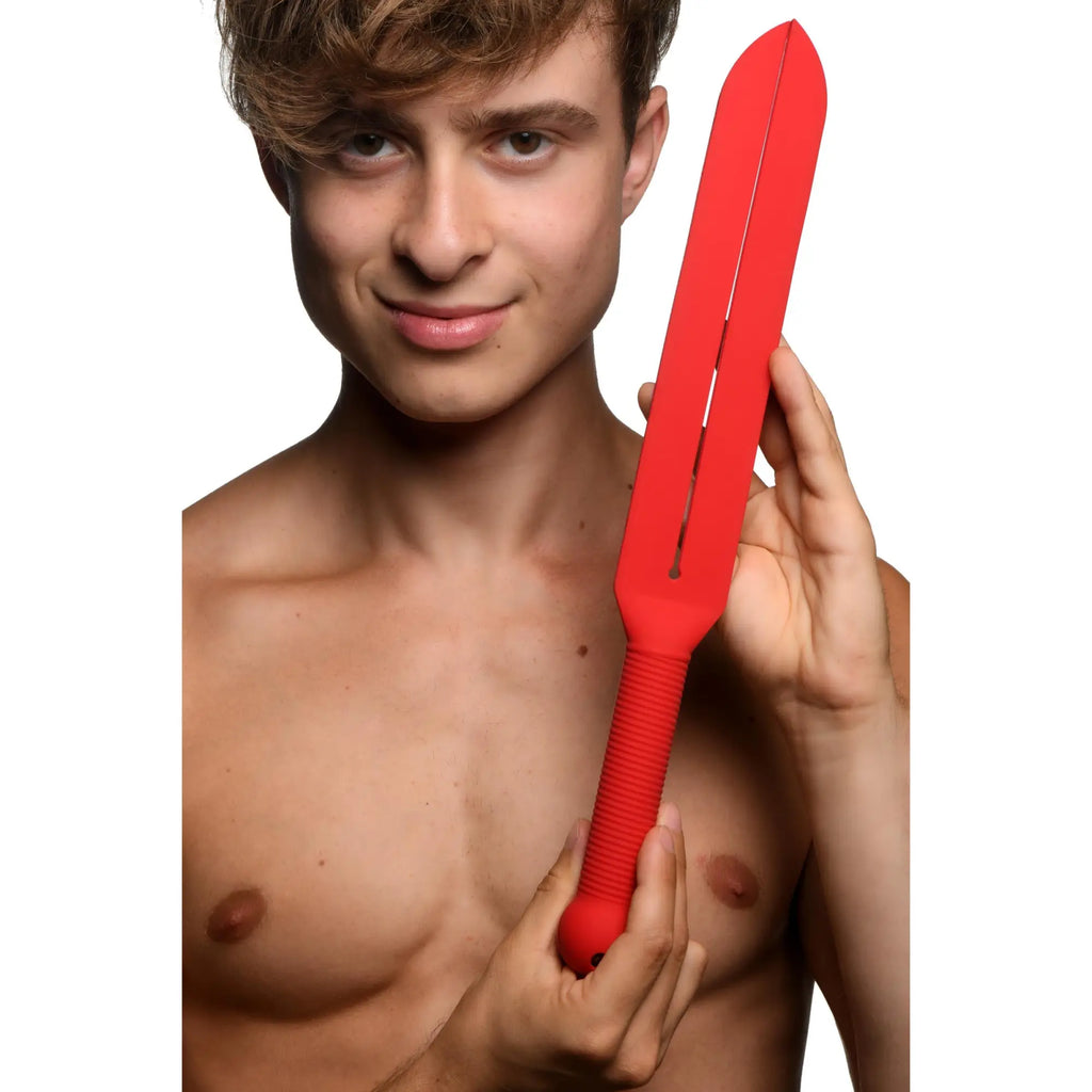 A person holds a red, long, slotted Master Series Stung Silicone Tawse paddle with a textured, ridged grip handle.