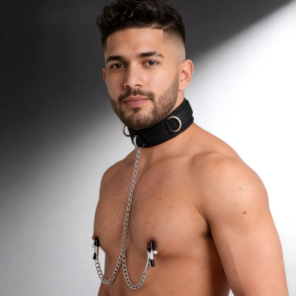Man wears a black Master Series submission collar connected by a silver chain to matching nipple clamps.