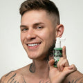 Smiling tattooed man holding a bottle of Master Series Suck It Throat Desensitizing Spray with green liquid.