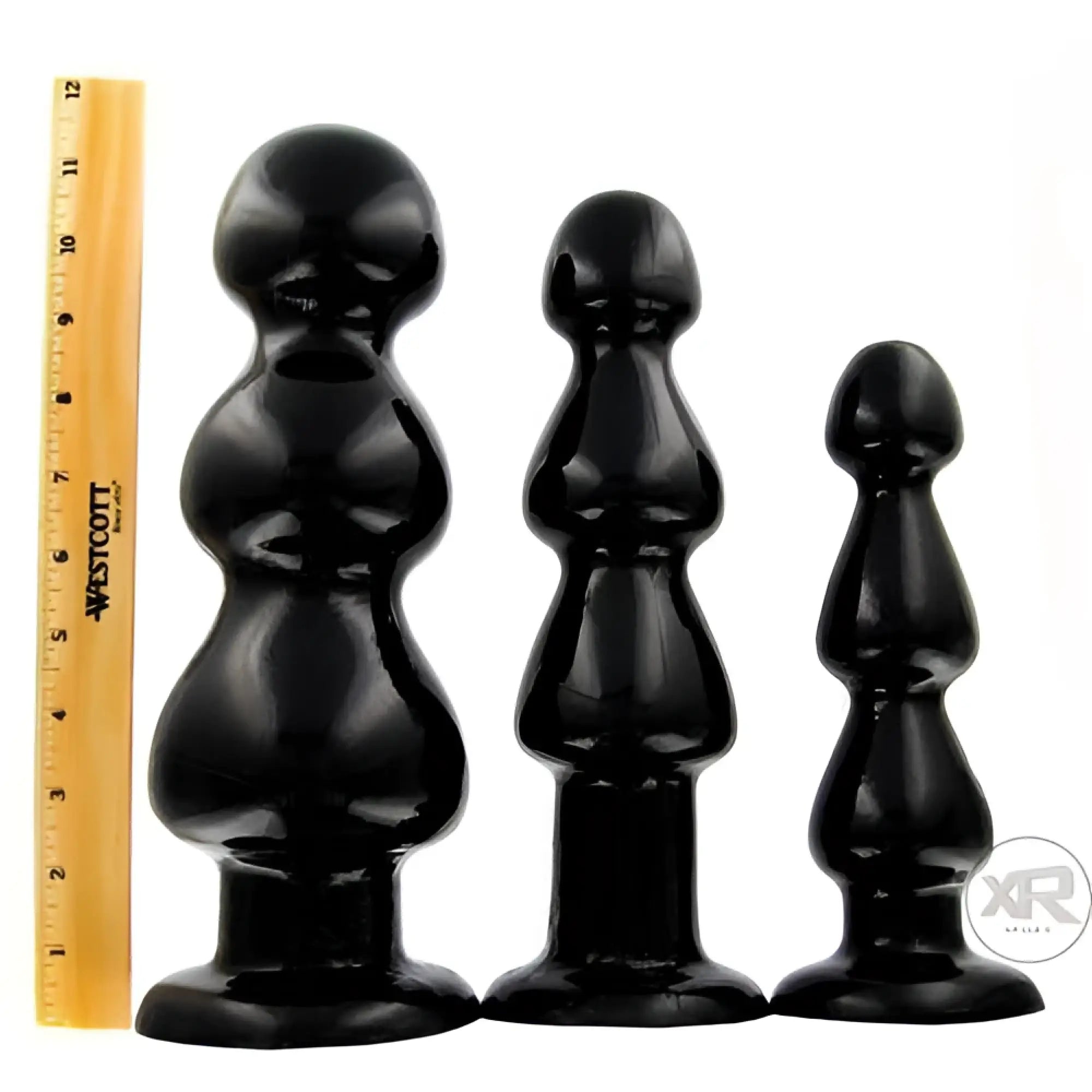 Three glossy black Master Series Three Bumps for Your Rump anal plugs in varying sizes next to a yellow ruler.