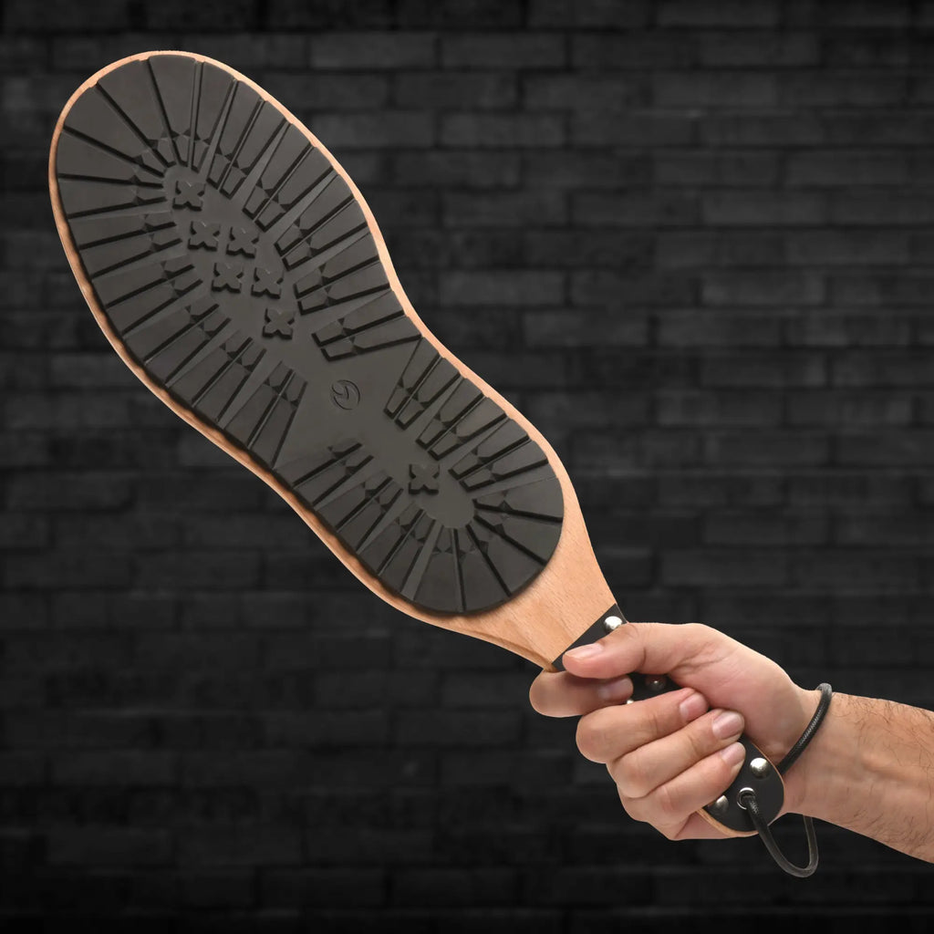 Hand holding a Master Series Tread Boot Paddle, featuring a wooden handle and a textured black rubber boot-sole face.