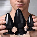 A man holds three glossy black, smooth, cone-shaped anal plugs of varying sizes from the Master Series 3-piece set.