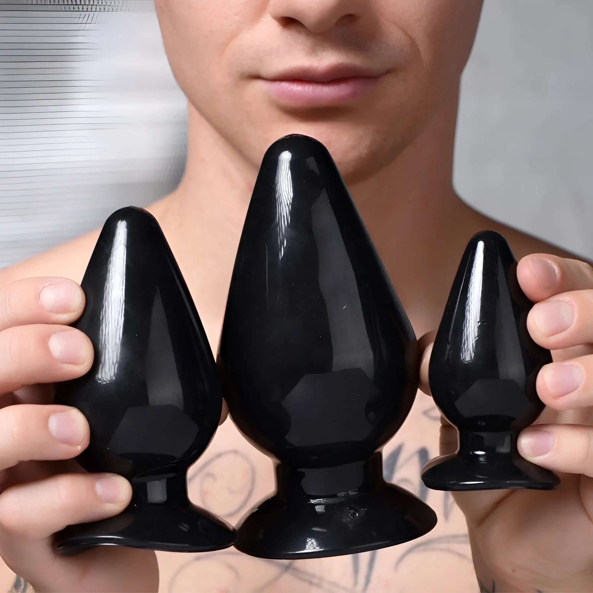 A man holds three glossy black, smooth, cone-shaped anal plugs of varying sizes from the Master Series 3-piece set.
