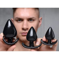 A person holding three glossy, black Master Series Triple Spades anal plugs of varying sizes with curved base grips.