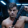 A man holds a black Master Series The Violator 13 Mode XL thruster, a realistic, textured dildo, against a blue background.