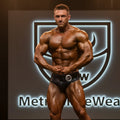 A muscular male bodybuilder wearing black shiny MetroMaleWear crocodile swim briefs with a silver number 3 badge