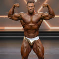 A muscular man flexing arms on stage wearing gold and white MetroMaleWear MetroMuscleWear competition briefs