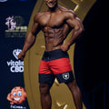 A muscular man wearing metroMalewear physique board short red with white bull logo on the left leg