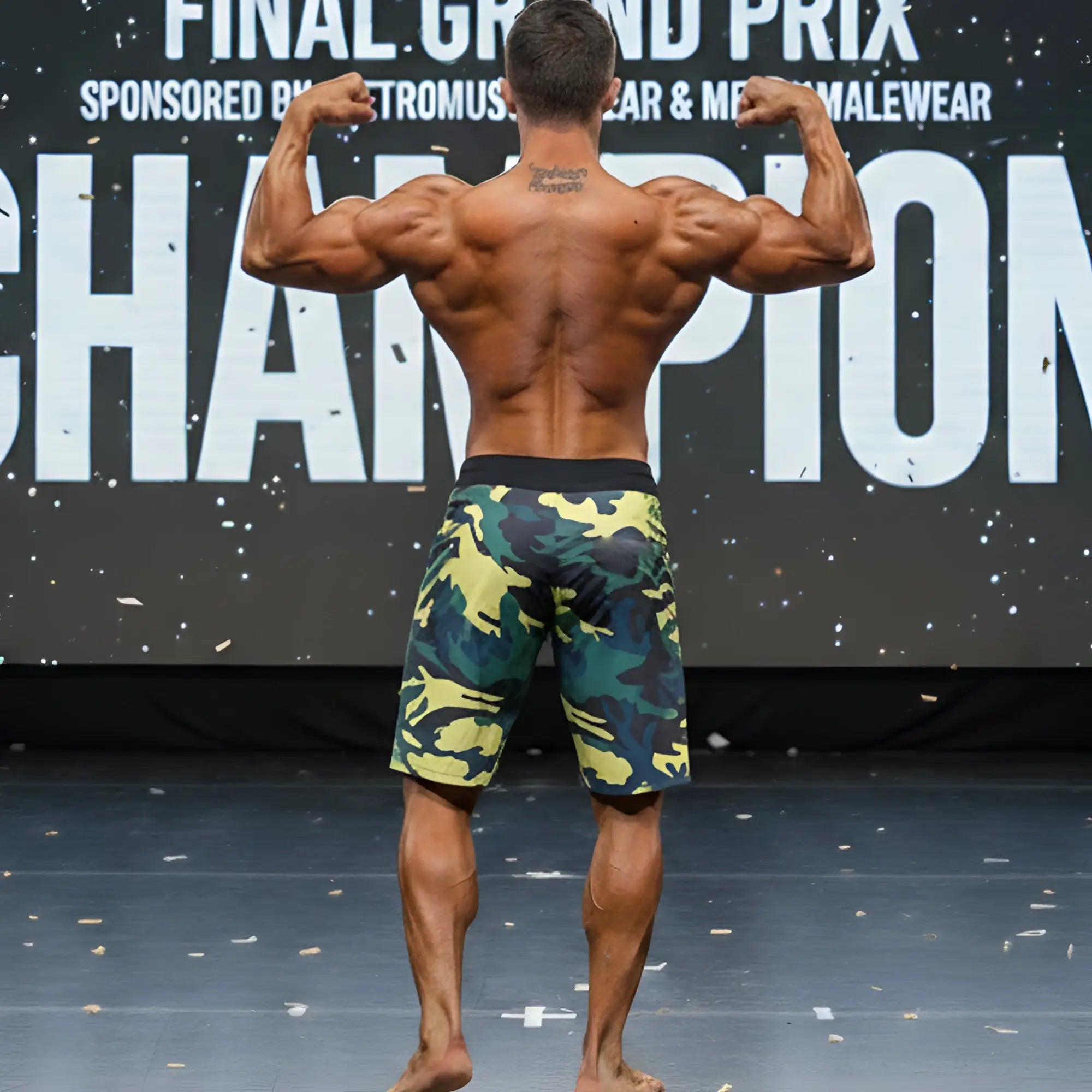 A muscular man stands on stage wearing metro male wear physique board short yellow camo with green and yellow patterns