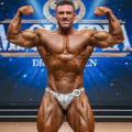 A muscular man wearing metroMalewear white and gray snake skin posing suit briefs with number 18 pinned on front