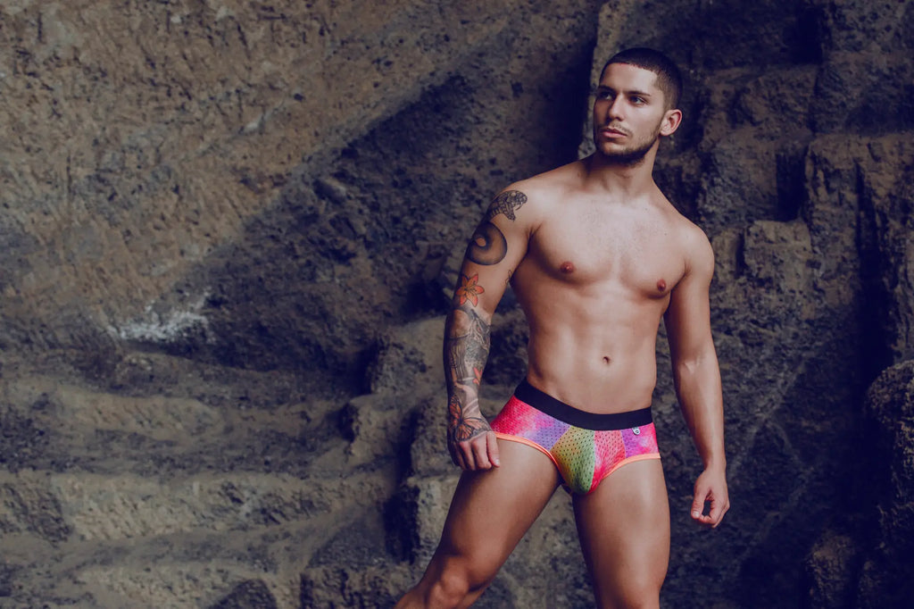 The man is wearing mob aero brief swim trunks in vibrant rainbow colors with a black waistband