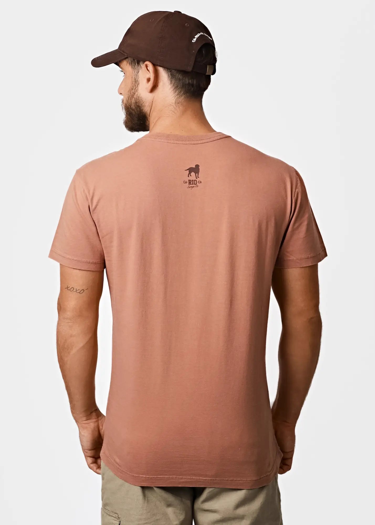Back of a dusty rose cotton men’s t-shirt featuring a small brown dog logo and text from praia do abrico tee rio famous nude beach
