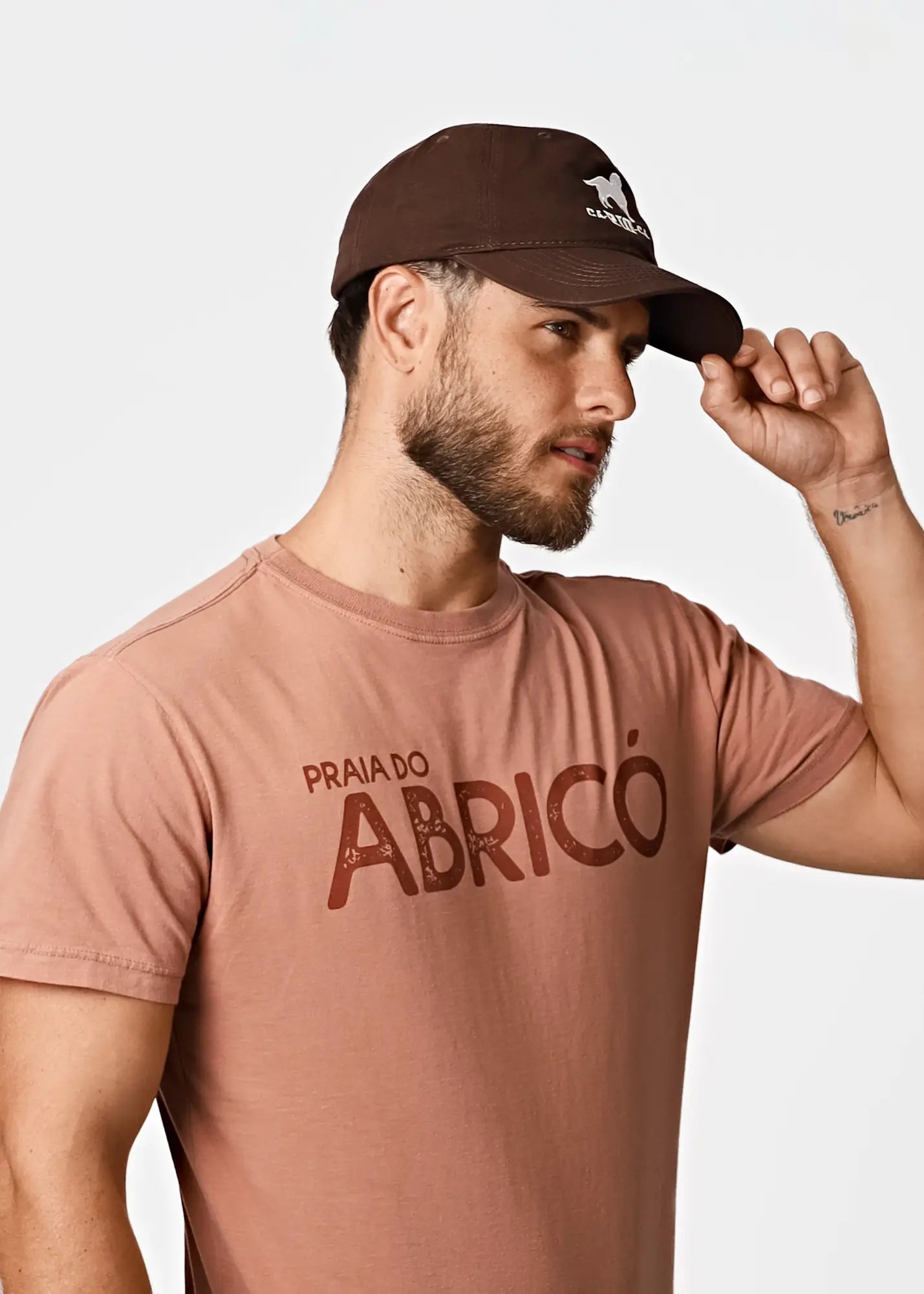 A man wearing a brown baseball cap with a white horse logo and carrasco embroidery with a praia do abrico tee