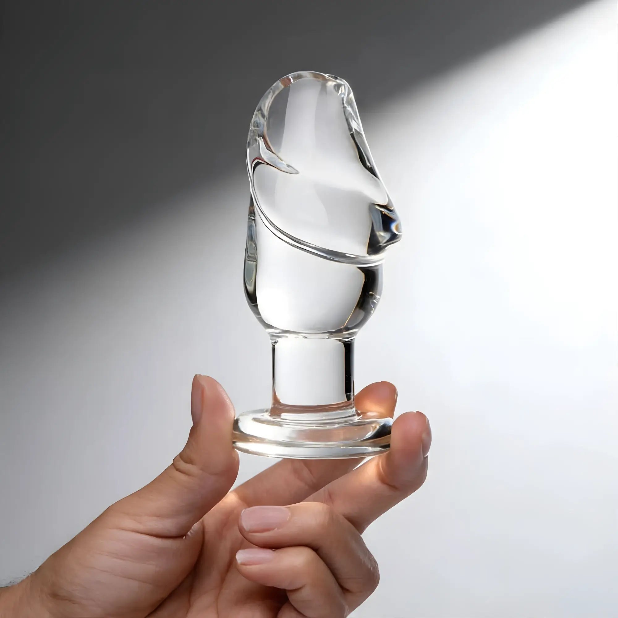 Clear glass Prisms Erotic cock head-shaped butt plug with a flat base, held by a hand against a gray backdrop.