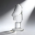 Prisms Erotic clear glass butt plug featuring a sculpted, cock-head shaped top on a round base against a light background.
