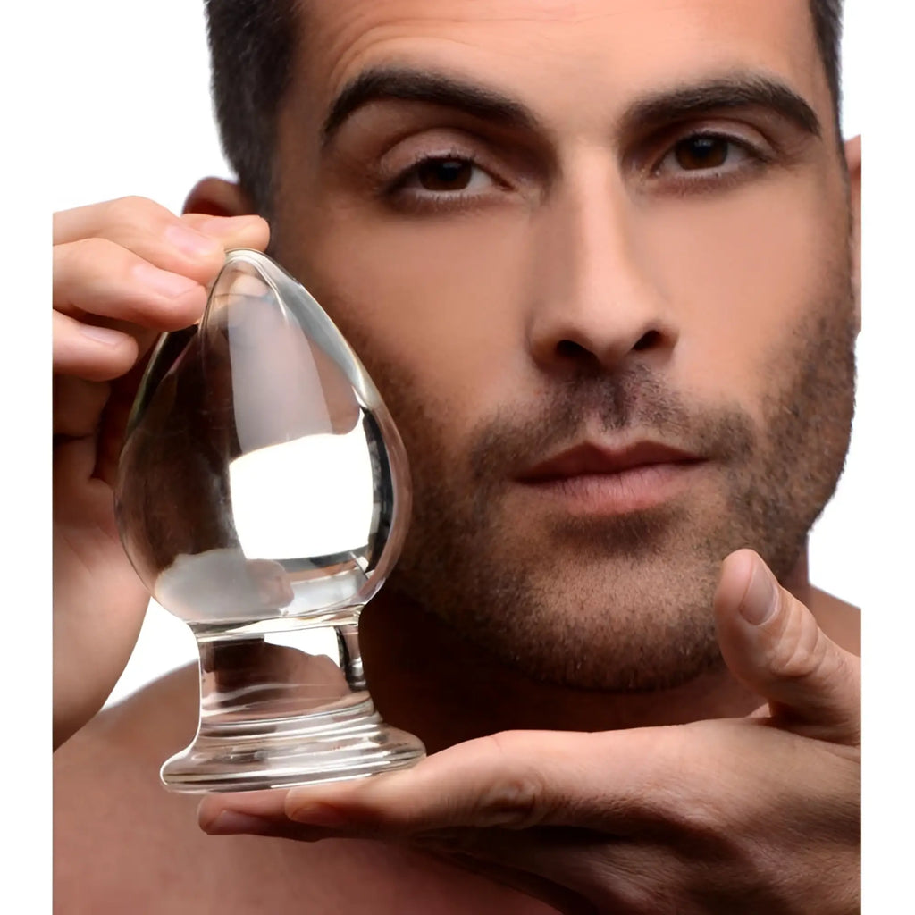 A man holds a clear, teardrop-shaped Prisms Erotic Glass wide butt plug against a white background.