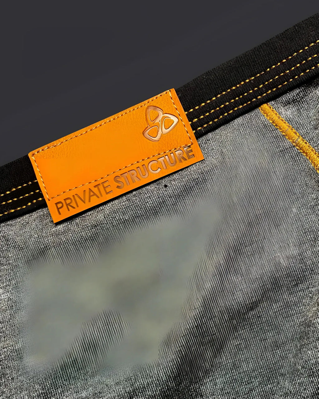 Orange leather brand tag with embossed logo and private structure text stitched on dark gray denim of barn boy mini brief
