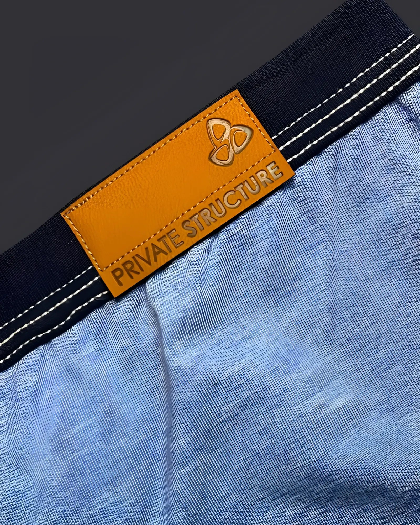 Orange leather brand tag with embossed logo and private structure text on dark blue denim waistband of private structure barn boy low rise trunk