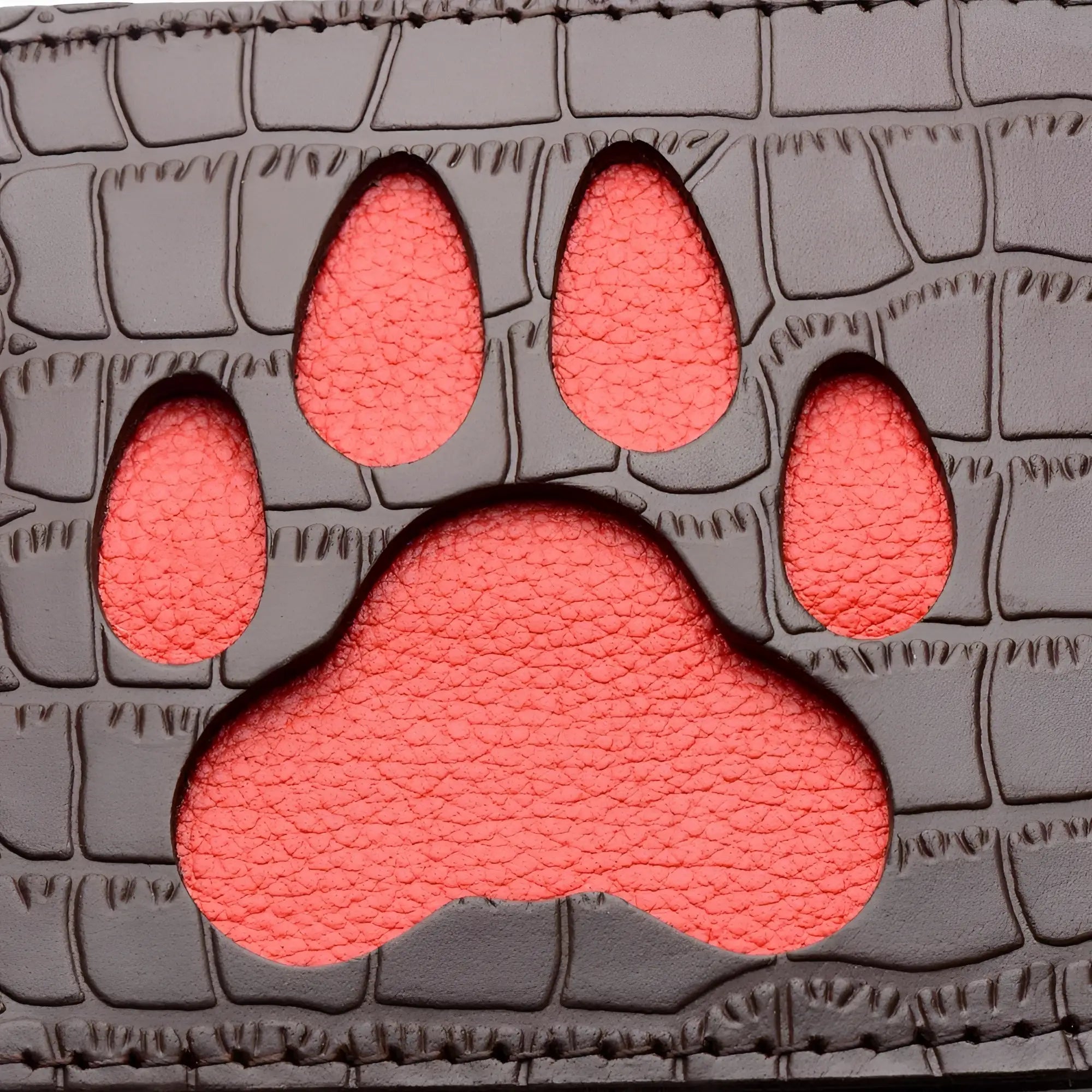 A coral-colored paw print cutout on a dark gray crocodile-textured leather surface of the puppy paw leather paddle