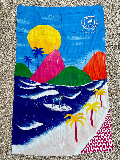 A vibrant Rio Aquarela canga de praia beach towel with colorful surf scene and dog surfing on waves in bright colors