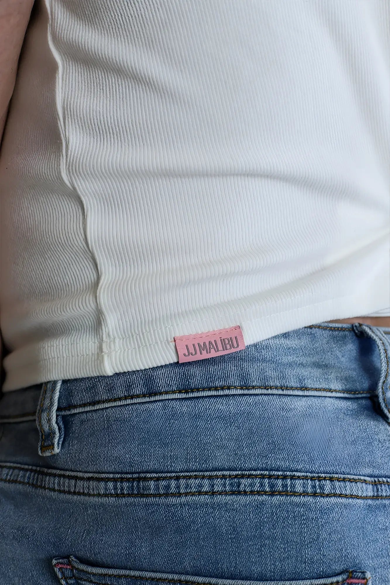 A pink jumalibu label stitched on white ribbed fabric of a scooped racer v tank top white