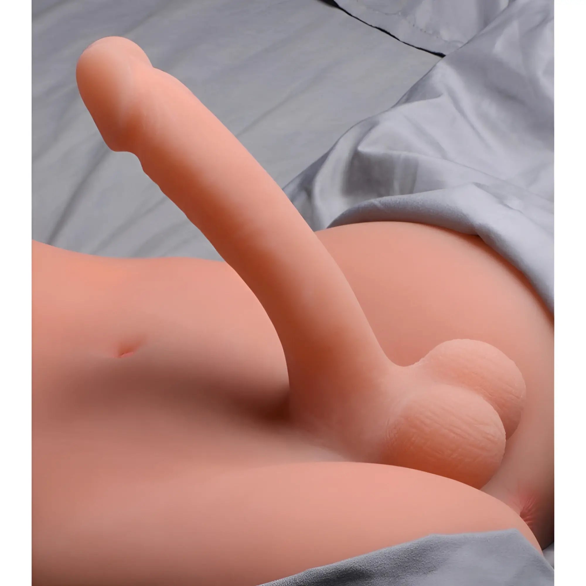 SexFlesh Trans Terri love doll showing a realistic, flesh-toned erect penis and scrotum on a grey bedsheet background.