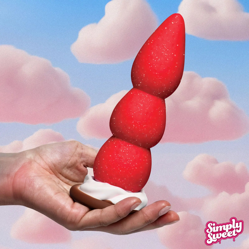 Hand holds a sparkly red, triple-bulb Simply Sweet Strawberry silicone dildo on a cream base against a cloudy sky.