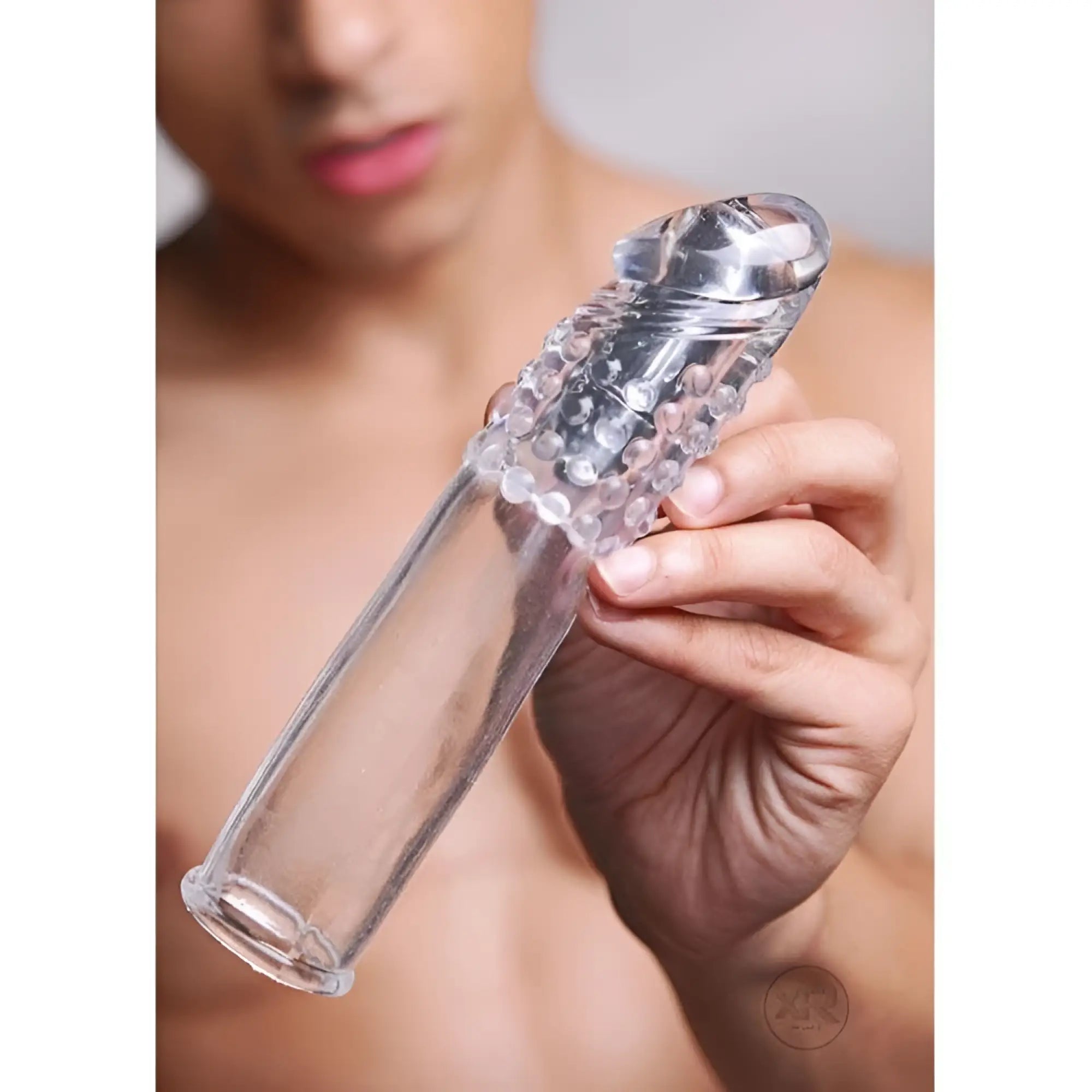 A hand holds a clear, textured Size Matters Clear Sensations vibrating penis enhancer.