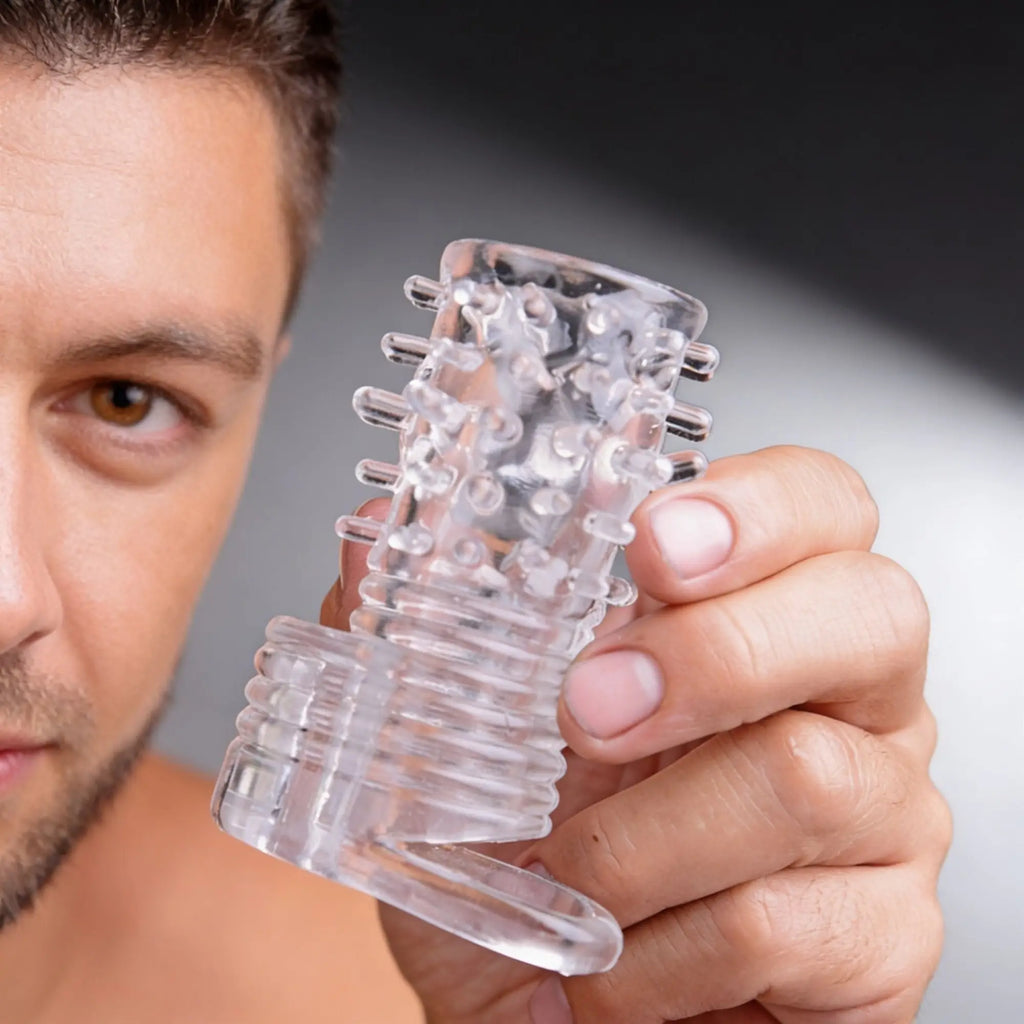 A man holds a clear, textured Size Matters Sensations vibrating erection sleeve featuring studs and a ribbed base.