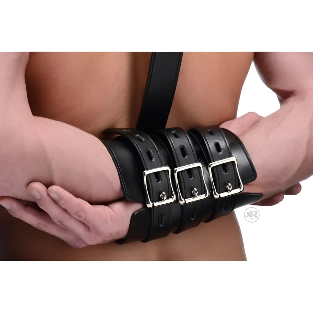 Black leather wrist restraint with three silver buckles, worn by a man with his hands held behind his back.