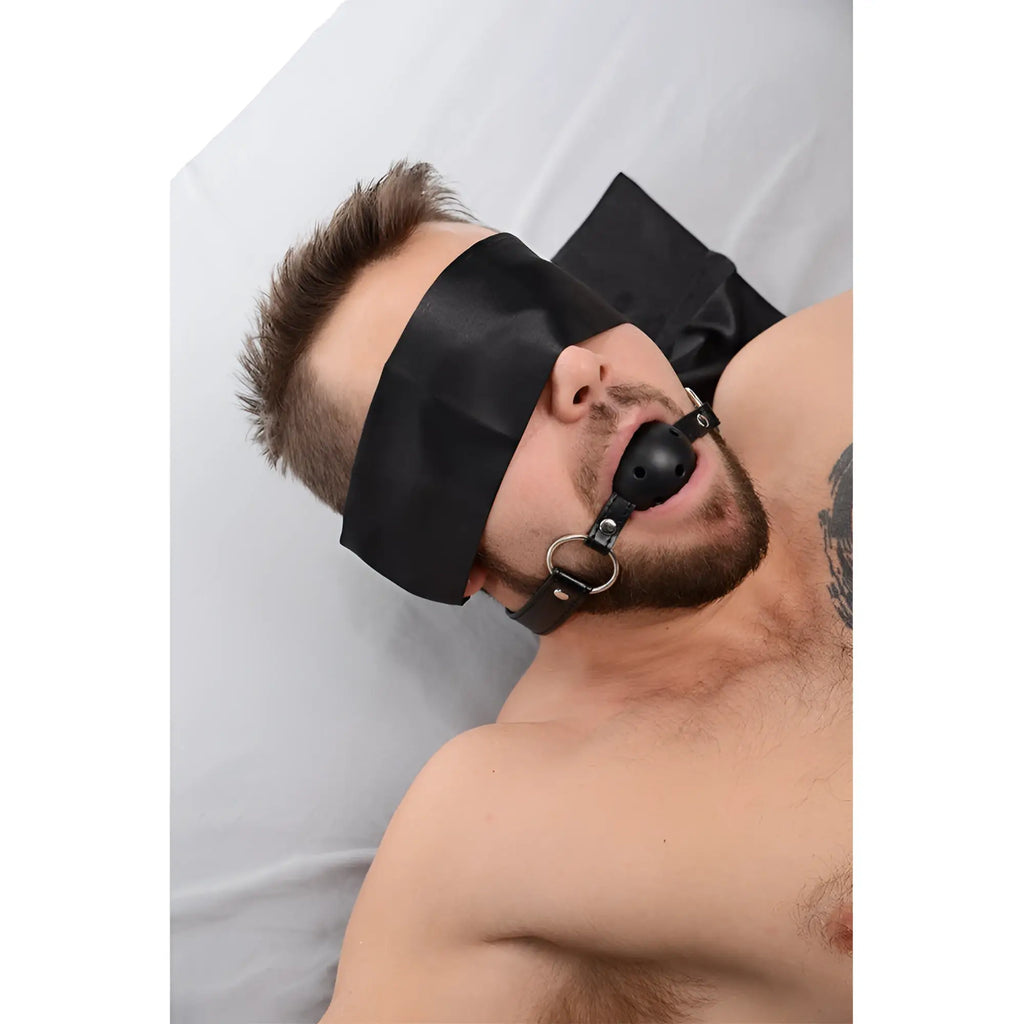 Strict Bed Restraint Bondage Kit: Person lying on bed wearing a black satin blindfold and black leather ball gag.