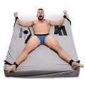 Strict Bed Restraint Bondage Kit: Man restrained on a grey bed with black wrist/ankle cuffs, gag, and accessories.